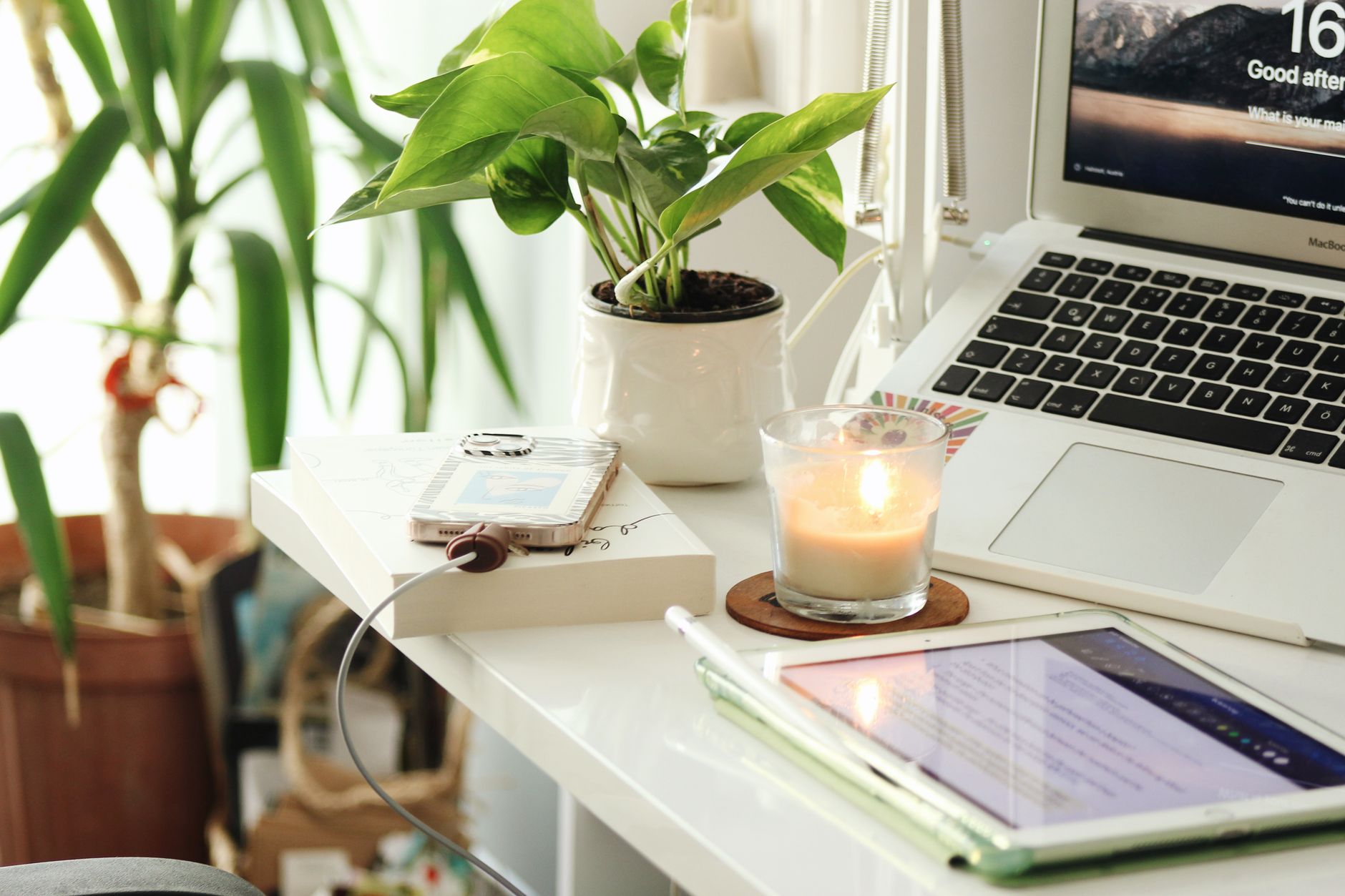 Secretária luminosa em casa com portátil, tablet, telemóvel, uma vela acesa e plantas verdes