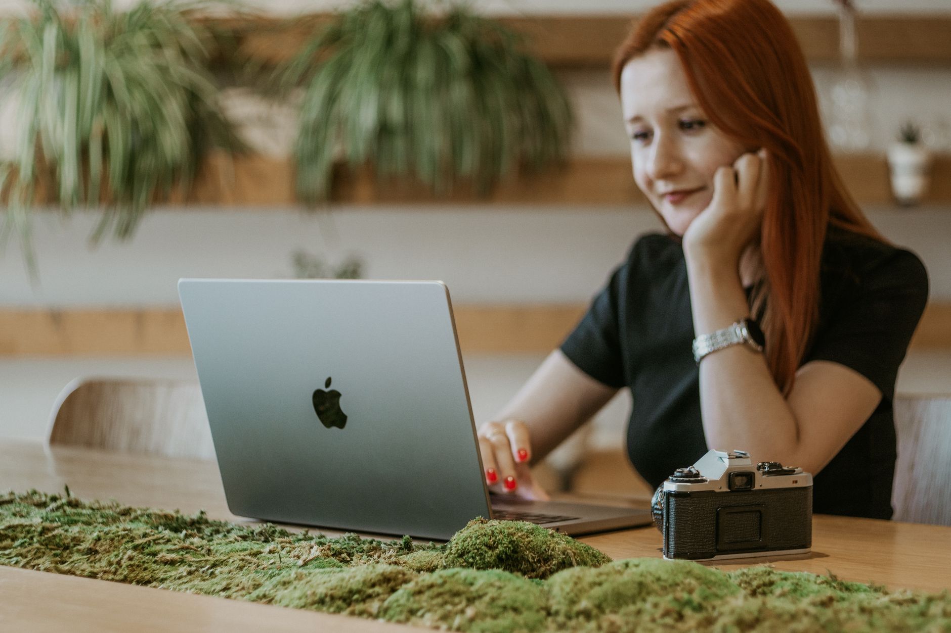 Mulher de cabelo ruivo sorridente a usar um portátil numa mesa de madeira, com um musgo decorativo e uma máquina fotográfica antiga